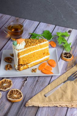 Slice of classic carrot cake with cream cheese frosting, topped with nuts and candied carrot, served with tea and walnuts on a rustic wood table