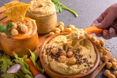 Koyu bir arka planda nohut, sebze ve diğer humus kavanozlarıyla çevrili ev yapımı kremalı humus kasesine mısır cipsi batırmak.