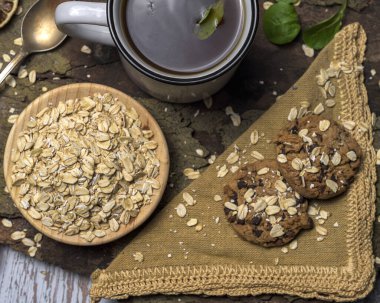 Kırsal ve sağlıklı kahvaltı tabağı çiğ yulaf, yeşil çay ve ahşap ve kumaş zemin üzerinde yulaf ezmeli çikolatalı kurabiye. Konforlu, doğal ve bitki temelli kavramlar için mükemmel.