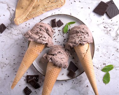 Üç çikolatalı dondurma külahı, üzerine serpiştirilmiş nane yaprakları ve mermer bir yüzey ve beyaz tabak.