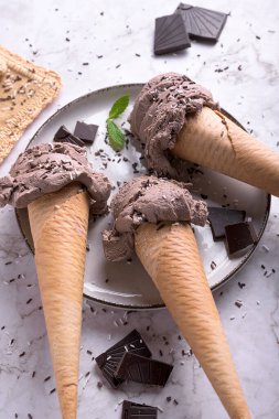 Üç çikolatalı dondurma külahı, üzerine serpiştirilmiş nane yaprakları ve mermer bir yüzey ve beyaz tabak.