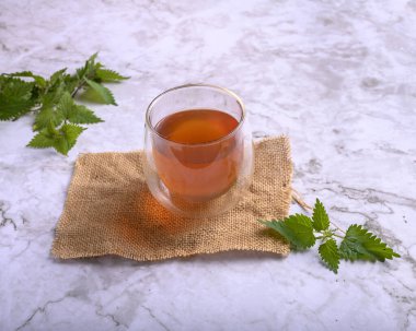Çuval bezi ve mermer yüzeyde çift duvar bardağında ısırgan otu çayı. Kavanozun etrafında taze ısırgan otu yaprakları, sağlıklı detoks içeceği ve doğal ilaç kavramı.