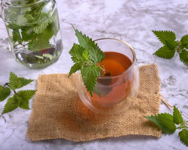 Çuval bezi ve mermer yüzeyde çift duvar bardağında ısırgan otu çayı. Kavanozun etrafında taze ısırgan otu yaprakları, sağlıklı detoks içeceği ve doğal ilaç kavramı.