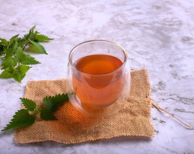 Çuval bezi ve mermer yüzeyde çift duvar bardağında ısırgan otu çayı. Kavanozun etrafında taze ısırgan otu yaprakları, sağlıklı detoks içeceği ve doğal ilaç kavramı.