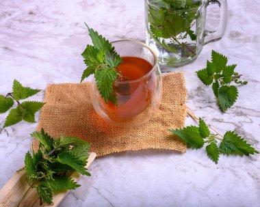 Çuval bezi ve mermer yüzeyde çift duvar bardağında ısırgan otu çayı. Kavanozun etrafında taze ısırgan otu yaprakları, sağlıklı detoks içeceği ve doğal ilaç kavramı.