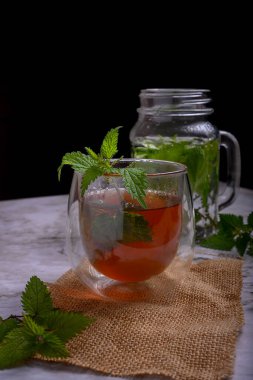 Isırgan otu çayı, çift cam bardakta, çuval bezi ve mermerde taze yapraklar, karamsar arka plan, dramatik ışıklandırma, bitkisel tıp ve sağlıklı yaşam tarzı konsepti, organik detoks içeceği.