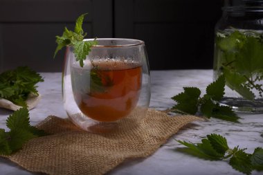 Isırgan otu çayı, çift cam bardakta, çuval bezi ve mermerde taze yapraklar, karamsar arka plan, dramatik ışıklandırma, bitkisel tıp ve sağlıklı yaşam tarzı konsepti, organik detoks içeceği.