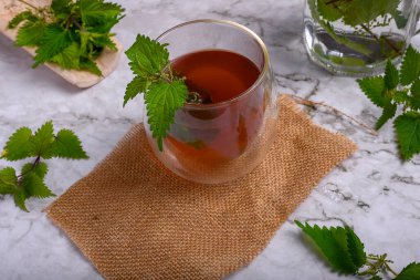 Çuval bezi ve mermer yüzeyde çift duvar bardağında ısırgan otu çayı. Kavanozun etrafında taze ısırgan otu yaprakları, sağlıklı detoks içeceği ve doğal ilaç kavramı.