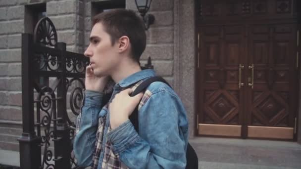 Молодий сучасник стоїть біля будинку на вулиці і розмовляє по телефону — стокове відео