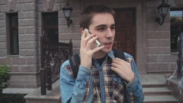 Joven hombre contemporáneo caminando en la calle cerca de la casa y hablando por teléfono — Vídeos de Stock