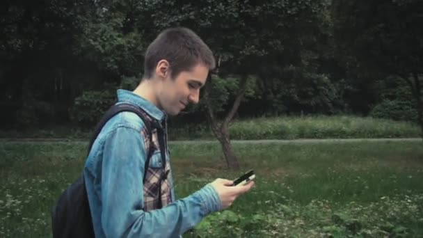 Jovem contemporâneo caminhando no parque e mensagens de texto em seu telefone usando o aplicativo no smartphone — Vídeo de Stock