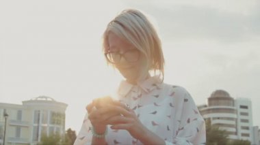 Onun smartphone Şehir Parkı manifatura app arka planda parlayan güneş ile kaydırma Internet tarama arkadaşları kullanan genç güzel kadın