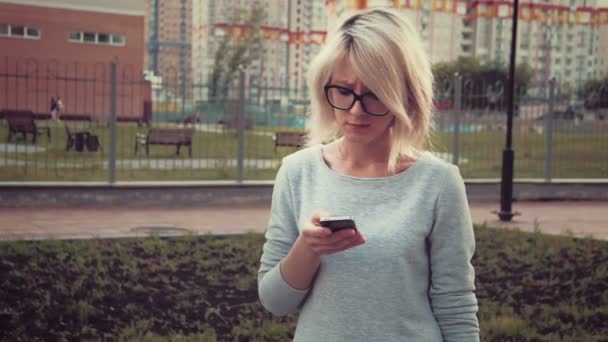 Грустная молодая женщина, стоящая в парке среди зданий, используя смартфон смс, грустит — стоковое видео