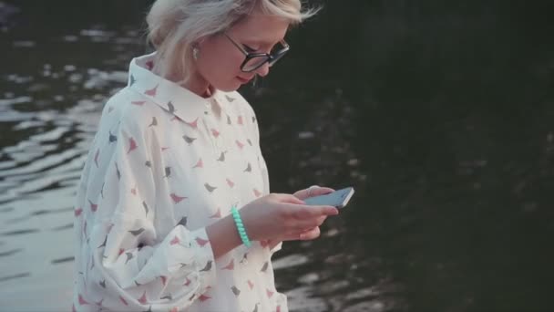 Νεαρή όμορφη γυναίκα, χρησιμοποιώντας smartphone της στο πάρκο της πόλης, κοντά στο η λίμνη γραπτών μηνυμάτων τους φίλους της ξεφυλλίσματος Διαδικτύου κύλιση εφαρμογή — Αρχείο Βίντεο