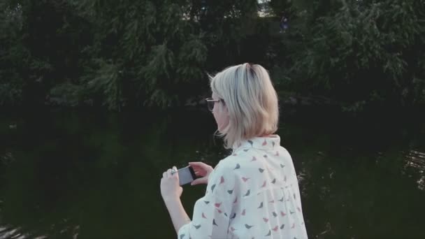 Joven hermosa mujer tomando fotos con su teléfono inteligente en el parque de la ciudad cerca del lago — Vídeos de Stock