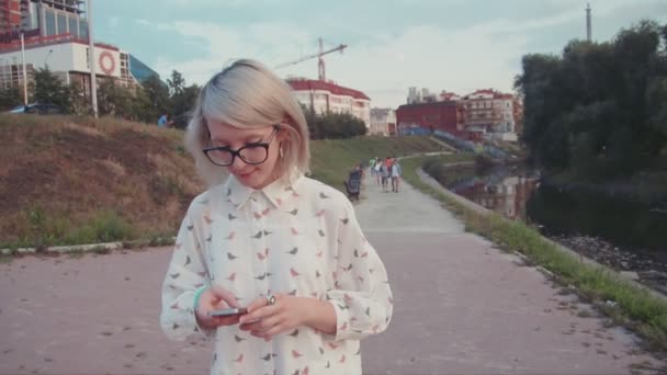 Νεαρή όμορφη γυναίκα που περπατά στο πάρκο κοντά στη λίμνη χρησιμοποιώντας το smartphone, γραπτών μηνυμάτων τους φίλους της, η περιήγηση στο internet, κύλιση εφαρμογή — Αρχείο Βίντεο