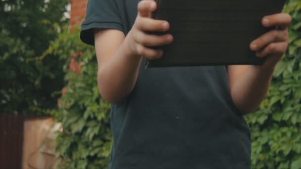 Mały chłopiec, grając w gry na jego ekran dotykowy tablet na zewnątrz przed domem pokryte zielenią — Wideo stockowe