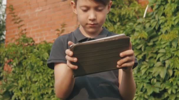 Kleine jongen spelen van spelletjes op zijn tablet touchscreen buitenshuis voor het huis bedekt met groen — Stockvideo