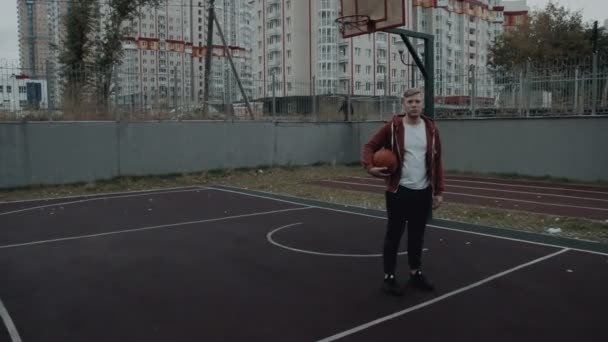 Joven wita una pelota de canasta mirando a la cámara al aire libre en la cancha — Vídeo de stock