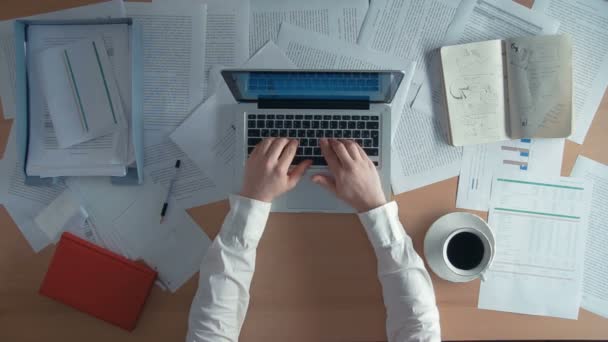 Geschäftsmann, der mit seinem Laptop am Schreibtisch sitzt, trinkt Kaffee und verschüttet ihn auf einem Blatt Papier, das er umgeben von viel Papierkram und Finanzberichten, Notizbüchern, wegwirft. — Stockvideo