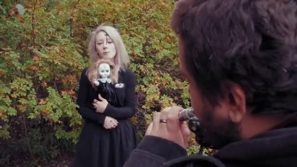 Tournage photo d'une jeune femme dans le caractère d'une fille effrayante au visage blanc avec une poupée — Video