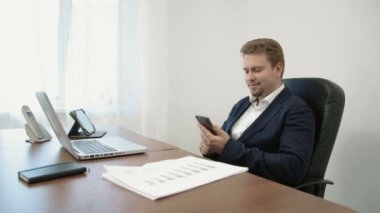 Dizüstü bilgisayar önünde ofisinde çalışan genç iş adamı. O iş app keşfetmek parmağıyla ekran dokunaklı onun smarphone kullanır.