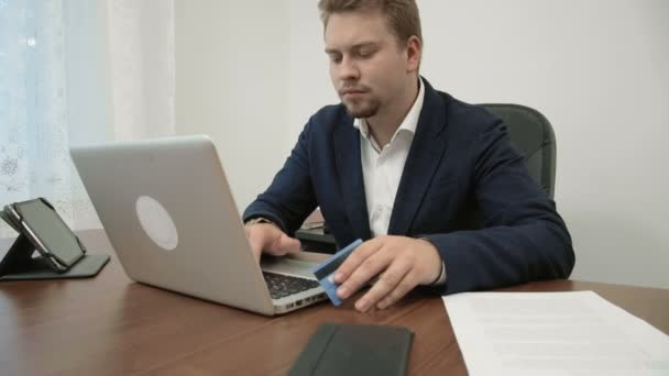 Junger Geschäftsmann macht Online-Einkäufe in seinem Büro, hält eine Kreditkarte in der Hand und tippt ihre Nummer in die Webseite ein — Stockvideo