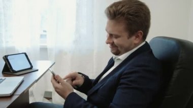 Dizüstü bilgisayar önünde ofisinde çalışan genç iş adamı. İş uygulaması keşfetmek parmağıyla ekran dokunaklı onun smarphone kullanır. O gülümseyen ve bakarak süre gülüyor
