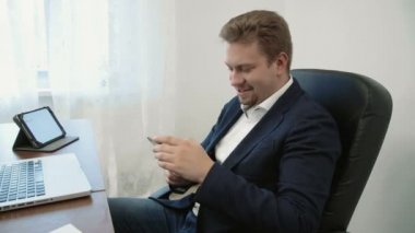 Dizüstü bilgisayar önünde ofisinde çalışan genç iş adamı. İş uygulaması keşfetmek parmağıyla ekran dokunaklı onun smarphone kullanır. O gülümseyen ve bakarak süre gülüyor