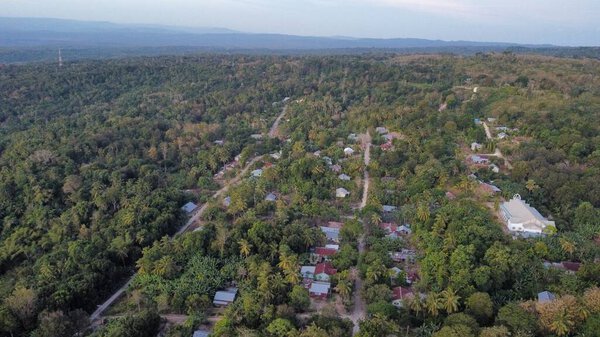 A drone's perspective reveals the delicate interplay between humanity and nature. Nestled in the foreground, a small, rustic farmstead with a corduroy pattern of crops rests at the foot of the towering, layered mountains.