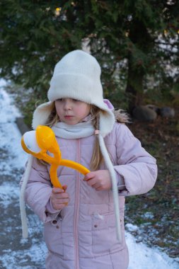 Pembe ceketli, beyaz şapkalı, karda oynayan, elinde turuncu bir kartopu makinesi olan genç bir kız. Arkaplanda parlak bir günde karlı zemin ve yeşil ağaçlar yer alıyor..