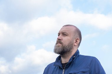 Sakallı bir adam dışarıda duruyor, bulutlarla dolu gökyüzüne bakıyor. Bugün açık bir gün ve o derin düşüncelere dalmış gibi görünüyor. Temiz havanın tadını çıkarıyor..