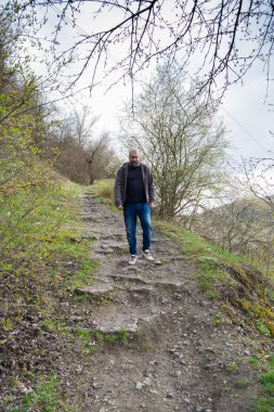 Bir adam patikayı çevreleyen çimler ve ağaçlarla kayalık bir patikada dikkatlice yürür. Bulutlu gökyüzü serin bir arkaplan ekler. Bahar yaprakları yukarıdaki dallarda görülebilir..
