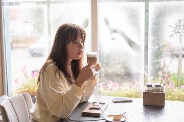 Kafedeki bir masada bir kadın oturuyor. Bir içki tutuyor ve sokağa bakarken düşünceli ve düşünceli görünüyor. Çiçekler pencereden görülebilir..