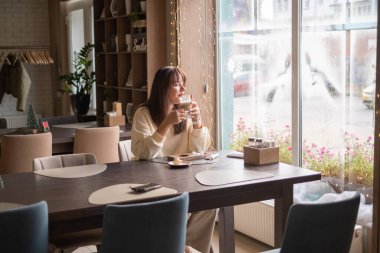 Kafedeki bir masada elinde fincanla oturan bir kadın. O, doğal ışıkla dolu büyük bir pencereden sokağı izler. Çiçekler dışarıda görünür..
