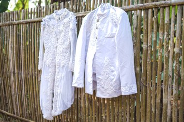 Gelinlik ve damadın takım elbisesi bambu bir çitte sergileniyor. Geleneksel bir açık hava düğünü için beyaz resmi kıyafetin detaylarını gözler önüne seriyor..