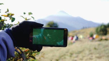 Elinde akıllı telefon bulunan kişi, açık havada dağ arkaplanı ve kamp çadırlarıyla gezme teknolojisi ve açık hava macerası öneren yeşil bir ekran sunuyor..