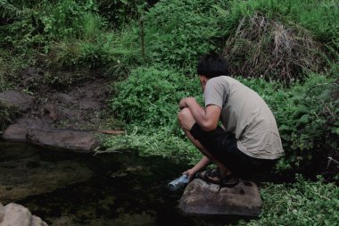 İnsan, yoğun yeşil doğal bitki örtüsüyle çevrili bir kayanın üzerinde çömelirken doğal bir dereden bir şişeye tatlı su toplar..
