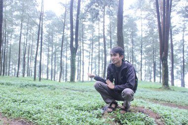 Genç Asyalı erkek gezgin yoğun bir çam ormanının yosunlu zeminine çömelir koyu renk giysiler giyerek ve düşünceli bir şekilde uzaklığa bakar..