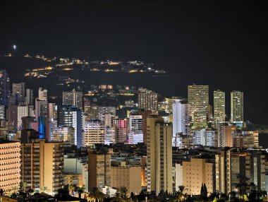 Gece vakti kıyı silueti parlıyor, feribotlar gece liman manzarasında parıldayan izler bırakıyor.