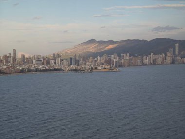 Huzurlu okyanus ve şehir manzarası, modern binalar ve günbatımının yer aldığı sakin kıyı şeridi