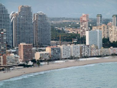 Gün ortası ufuk çizgisi manzarası, apartman blokları ve nazik okyanus dalgalarıyla güneş ışığı kıyısı.