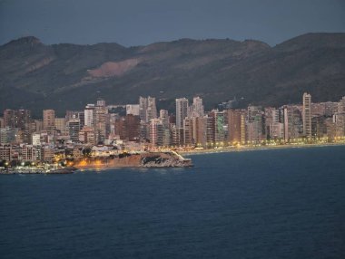 Canlı ışıklarla kıyı silüeti, hareketli liman ve dağ üzerinde alacakaranlık aydınlığı
