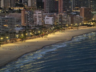 Alacakaranlıkta sakin deniz manzarası. Parlayan sokak lambaları ve boş güneşliklerle huzur yaratıyor.