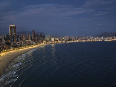 Akşam deniz kenarında parıldayan yürüyüş yolu, alacakaranlıkta hafif ışıklı gezinti güvertesi ve hafif dalgalarla