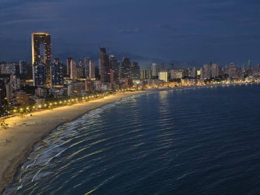 Aydınlatılmış kıyı şeridi olan şehir manzarası, ışıldayan sahil ve kentsel ufuk çizgisinin yer aldığı engin gece manzarası