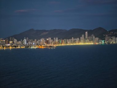 Uzak dağlarla kıyı boyunca ışıklar, şehir kıyı şeridi dağ manzaralarıyla parıldıyor.