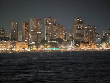 Canlı şehir silueti, gece boyunca huzurlu kıyı yüzeyi üzerinde yansıyan parlak pencerelerle.