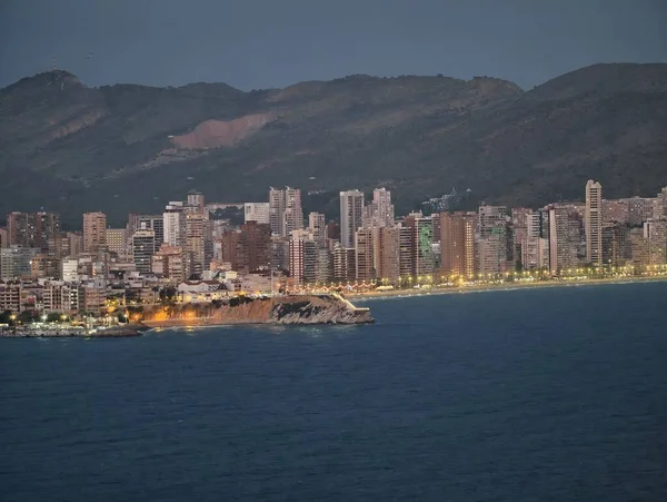 Canlı ışıklarla kıyı silüeti, hareketli liman ve dağ üzerinde alacakaranlık aydınlığı