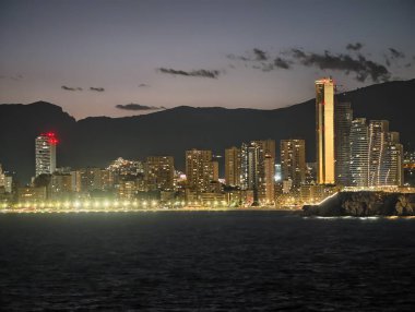 Dalgalarla dolu parlak kıyı şeridi, gece şehir silüeti ve canlı plaj ışıkları çarpıcı yaratıyor.